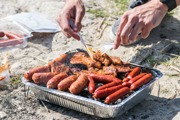 person holds cutlery, cooking meat on small foil tray disposable bbq on sandy ground