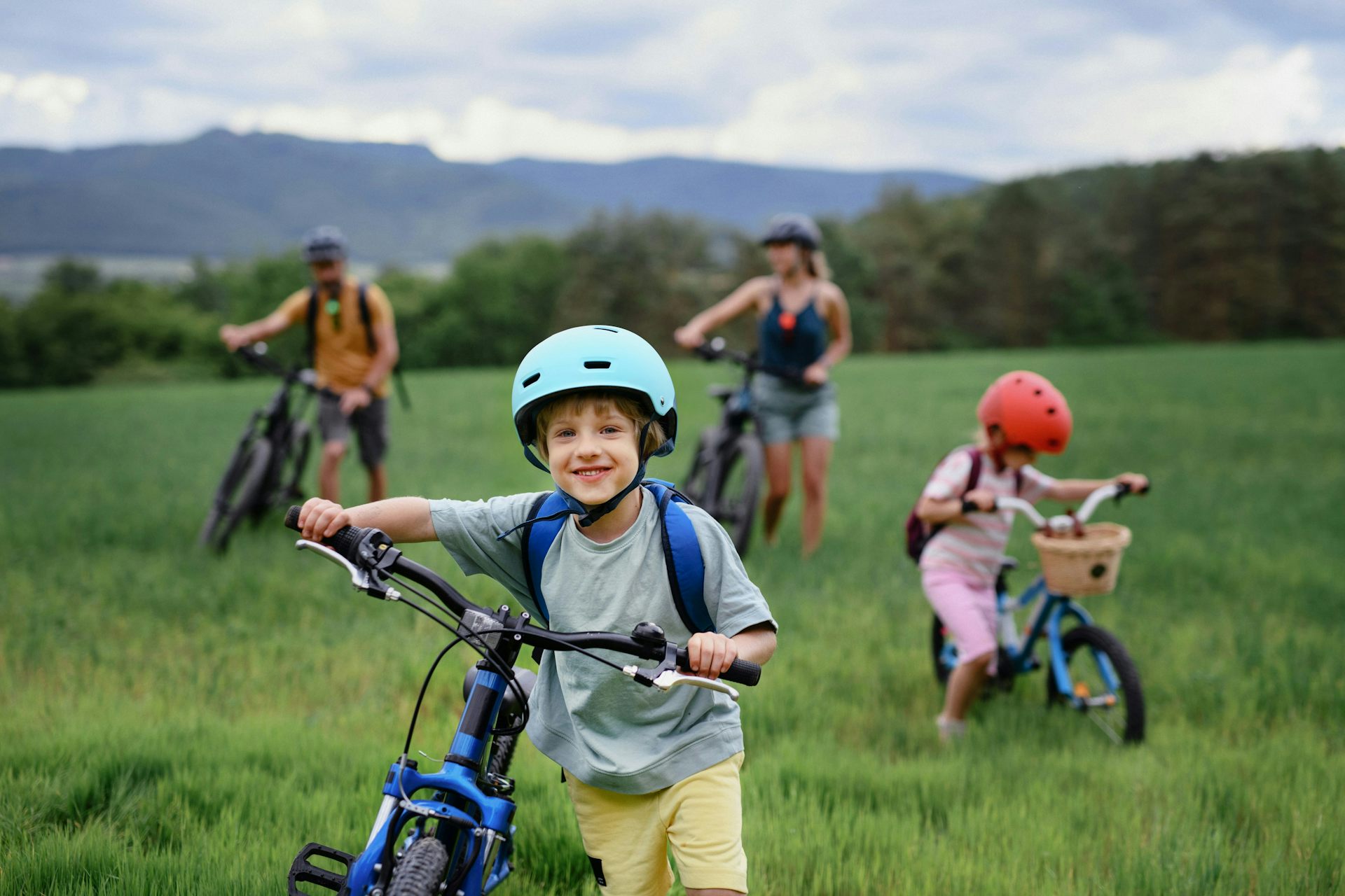 A child in a blue bike helmet with a blue bicycle in the foreground, with two adults on bikes and another child in a red helmet on a bike in the background in a grassy area