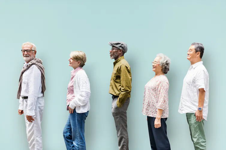 Five older adults standing in profile against a pale blue wall