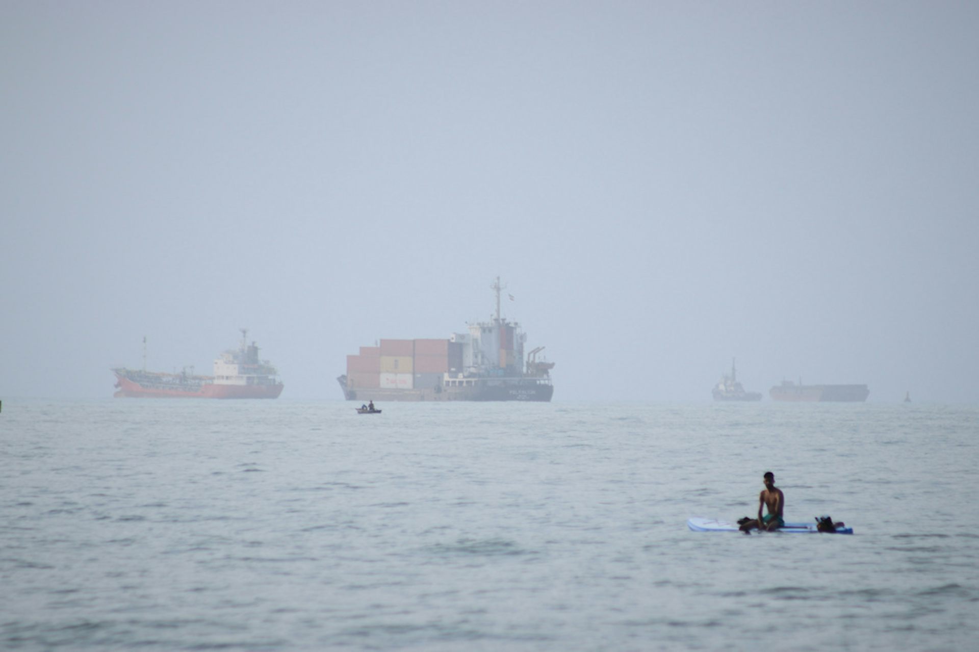 A man sits on a surfboard with container ships on the horizon.