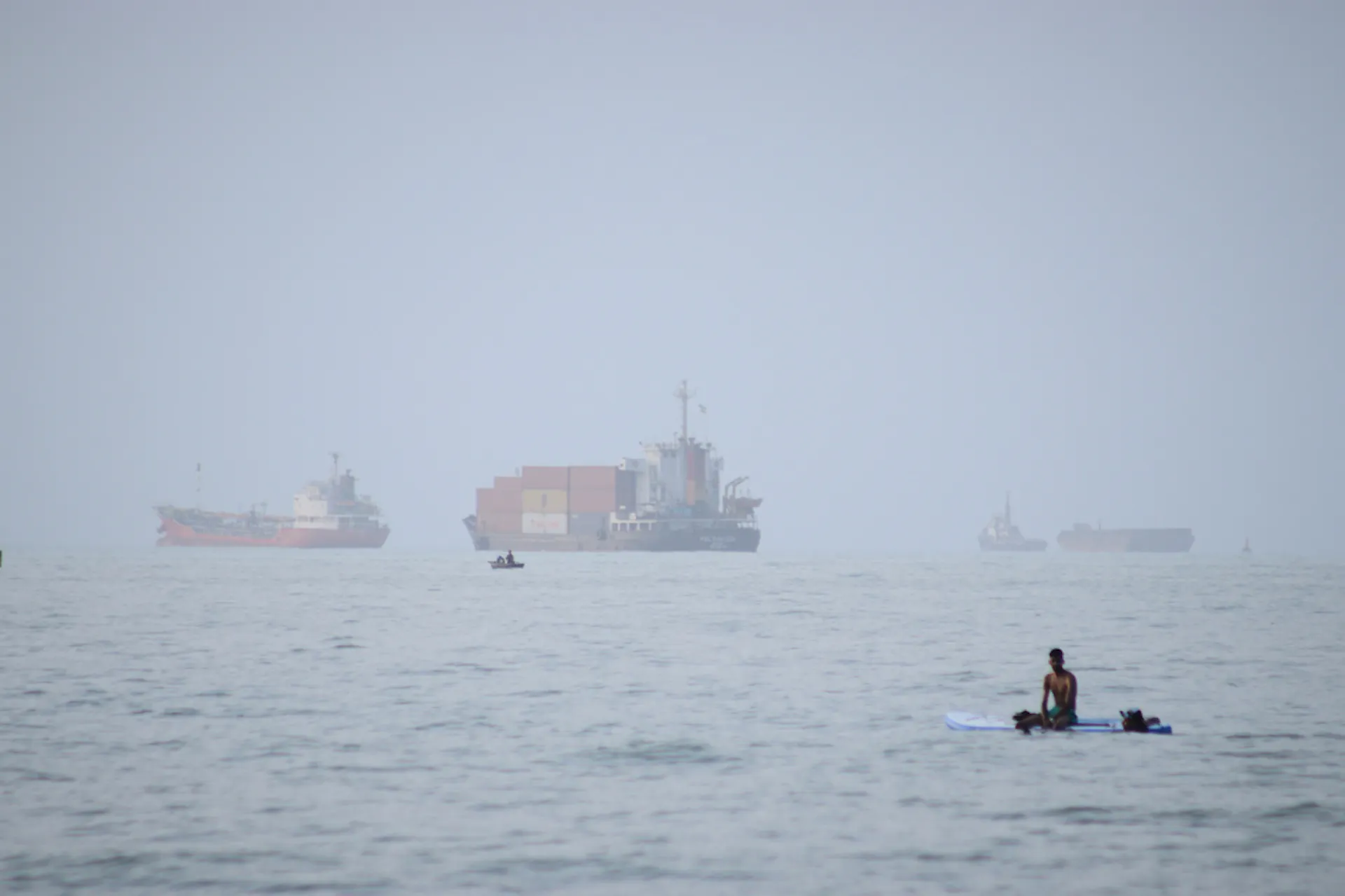 A man sits on a surfboard with container ships on the horizon.