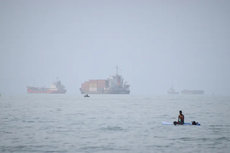 A man sits on a surfboard with container ships on the horizon.