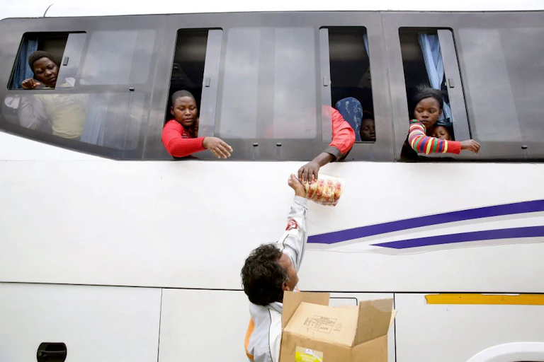 People looking out of bus windows as a person hands a loaf of bread up to them