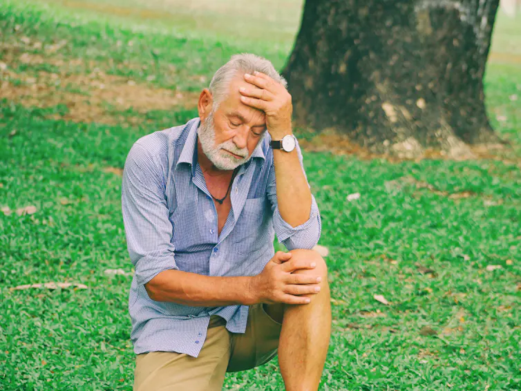 An elderly man crouches on the ground, holding his knee and head in pain.