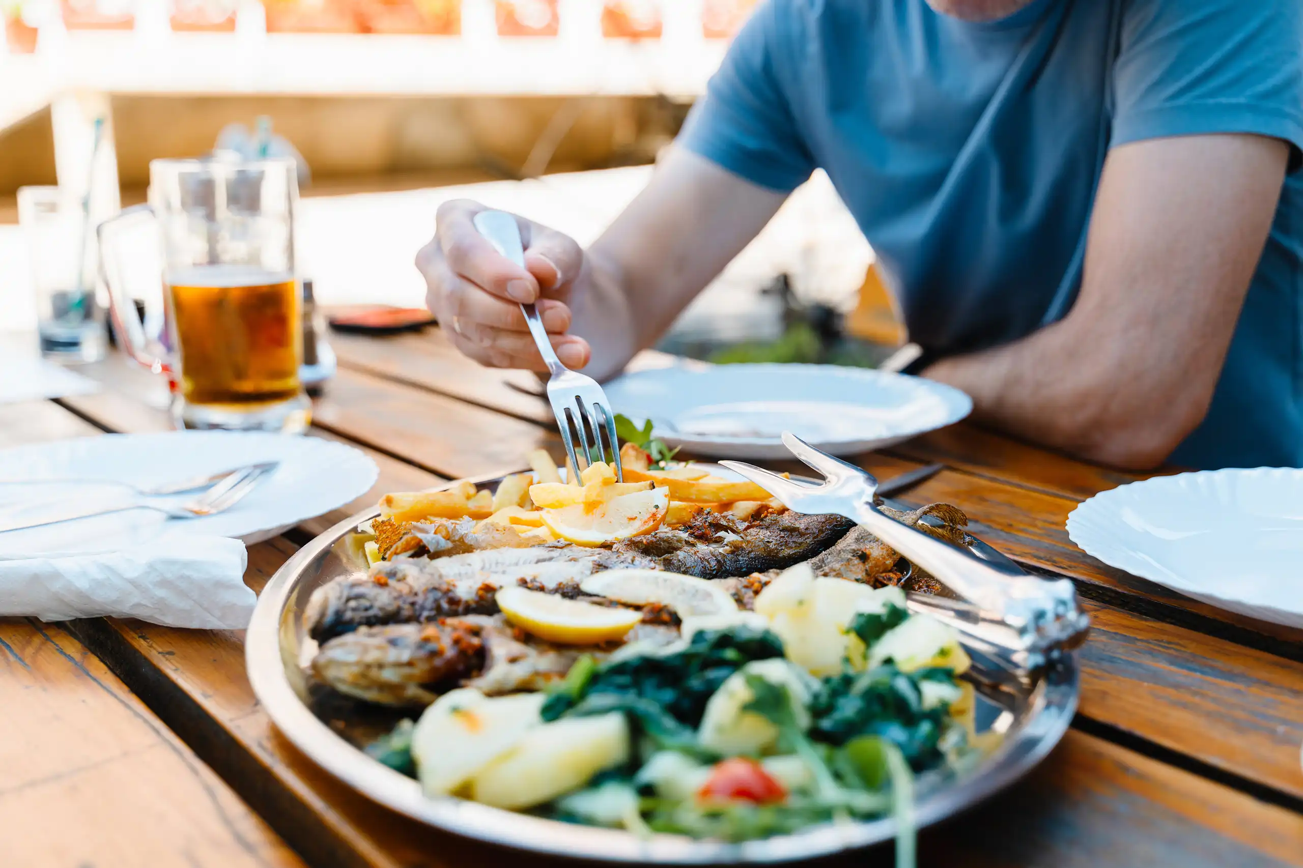 Une personne pique avec sa fourchette des frites dans une assiette, alors qu'elle mange sur une table en plein air.