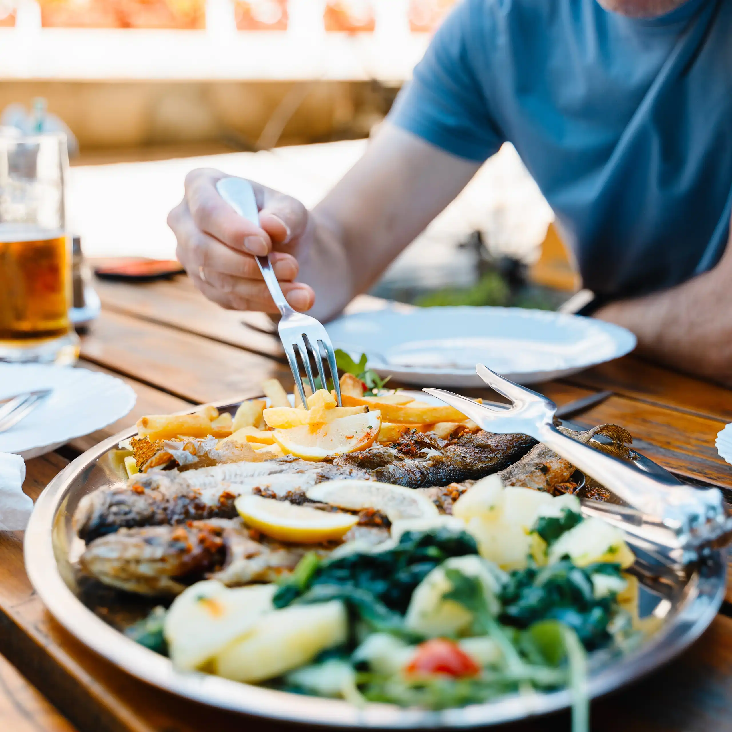 Une personne pique avec sa fourchette des frites dans une assiette, alors qu'elle mange sur une table en plein air.