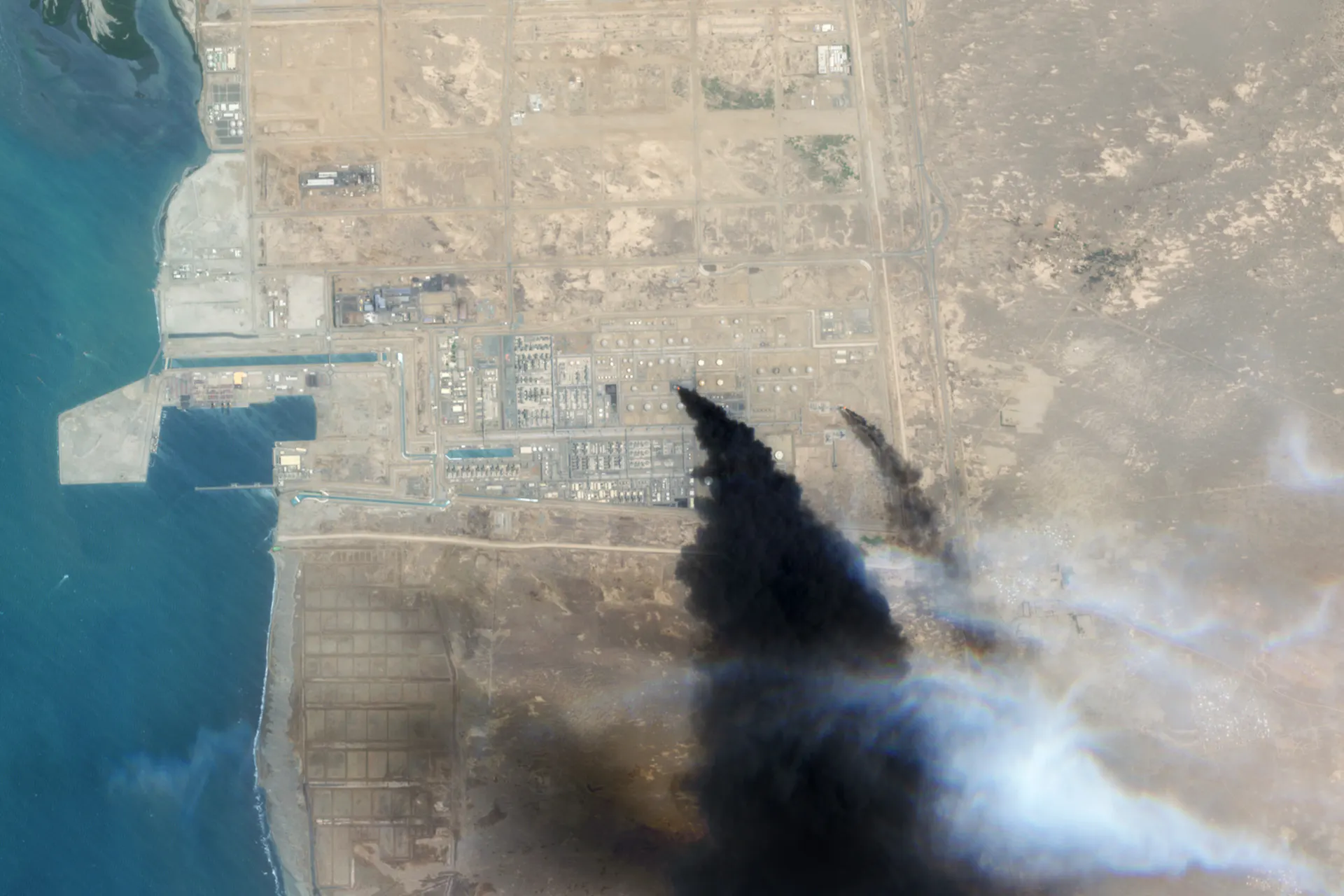 A satellite image shows massive plumes of black smoke rising from an industrial area along a coastline.