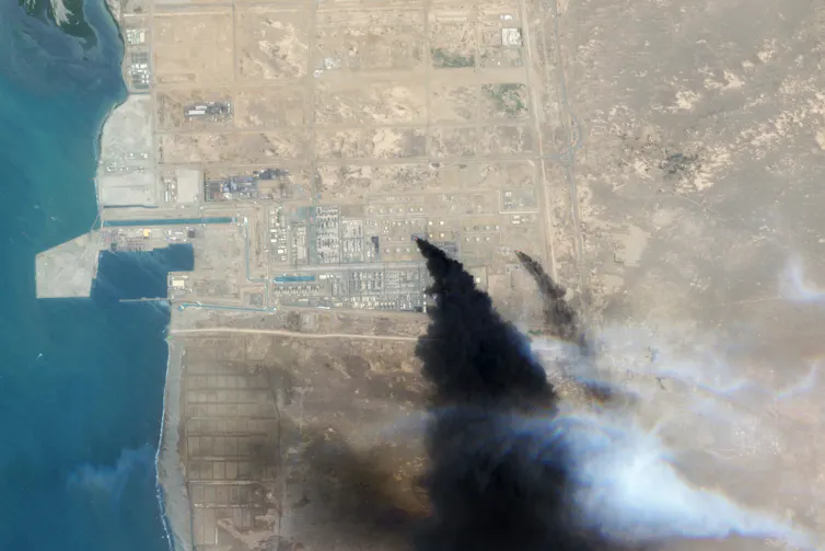 A satellite image shows massive plumes of black smoke rising from an industrial area along a coastline.