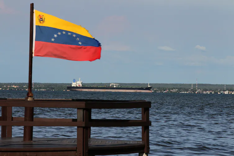 A white, blue and red flag flies in the foreground with a large tanker ship floating in the distance.