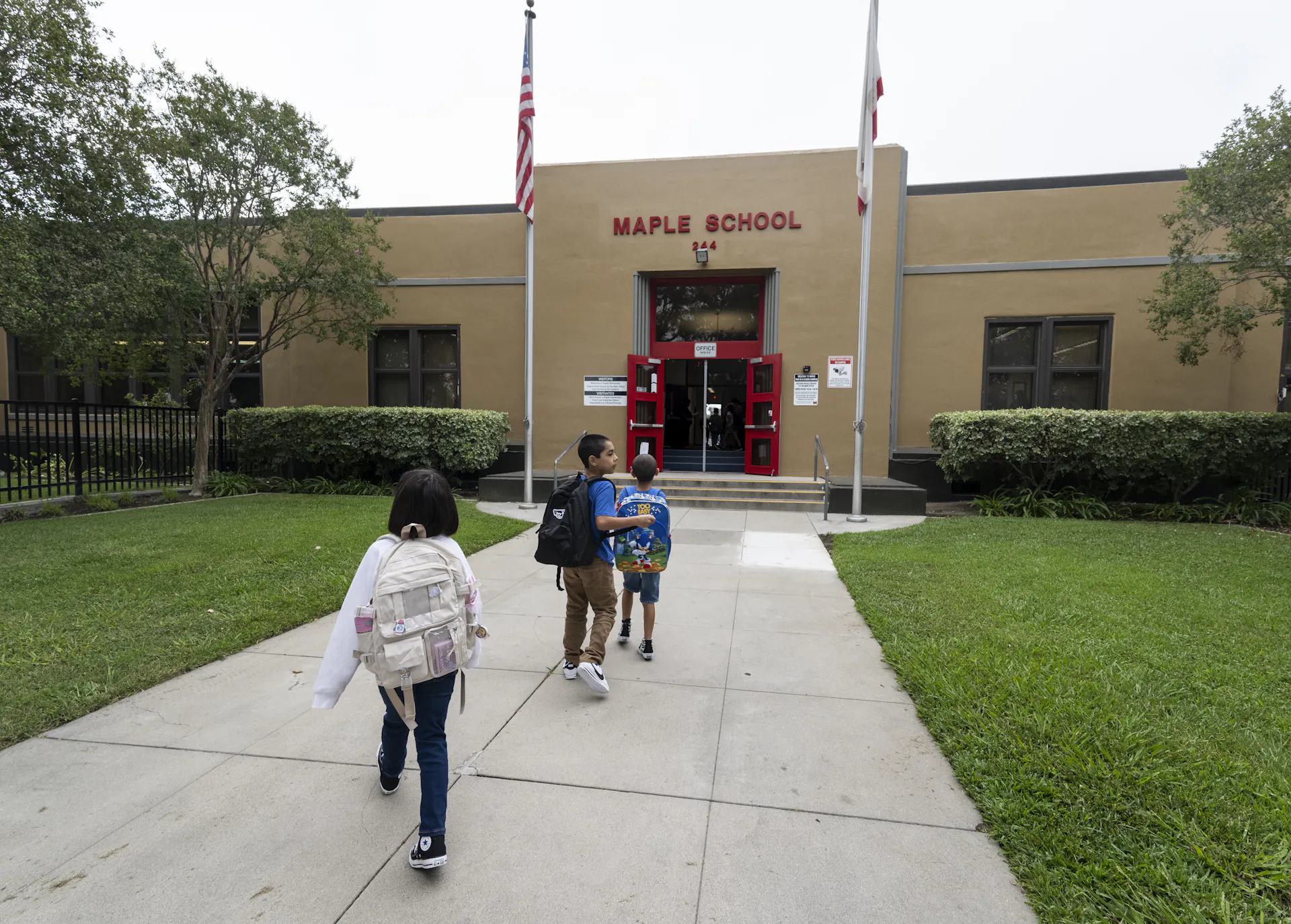 Three children walk on a sidewalk toward a brown building that has two flags raised outside of it.