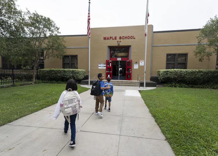 Three children walk on a sidewalk toward a brown building that has two flags raised outside of it.