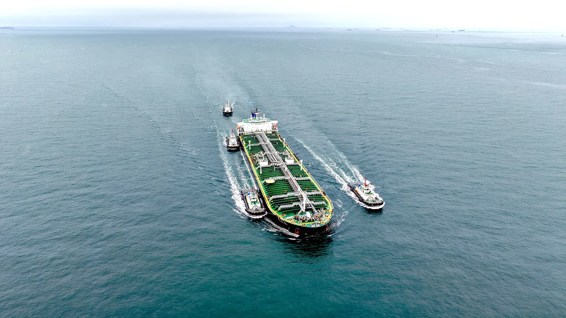 A large tanker ship sails across a body of water with multiple smaller boats alongside and nearby.