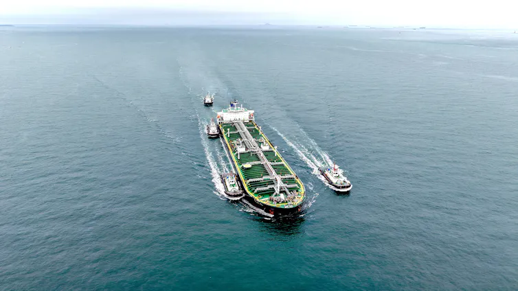 A large tanker ship sails across a body of water with multiple smaller boats alongside and nearby.