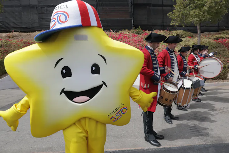 A person dresse in a yellow and white star costume poses next to a drum line decked out in colonial-era attire.