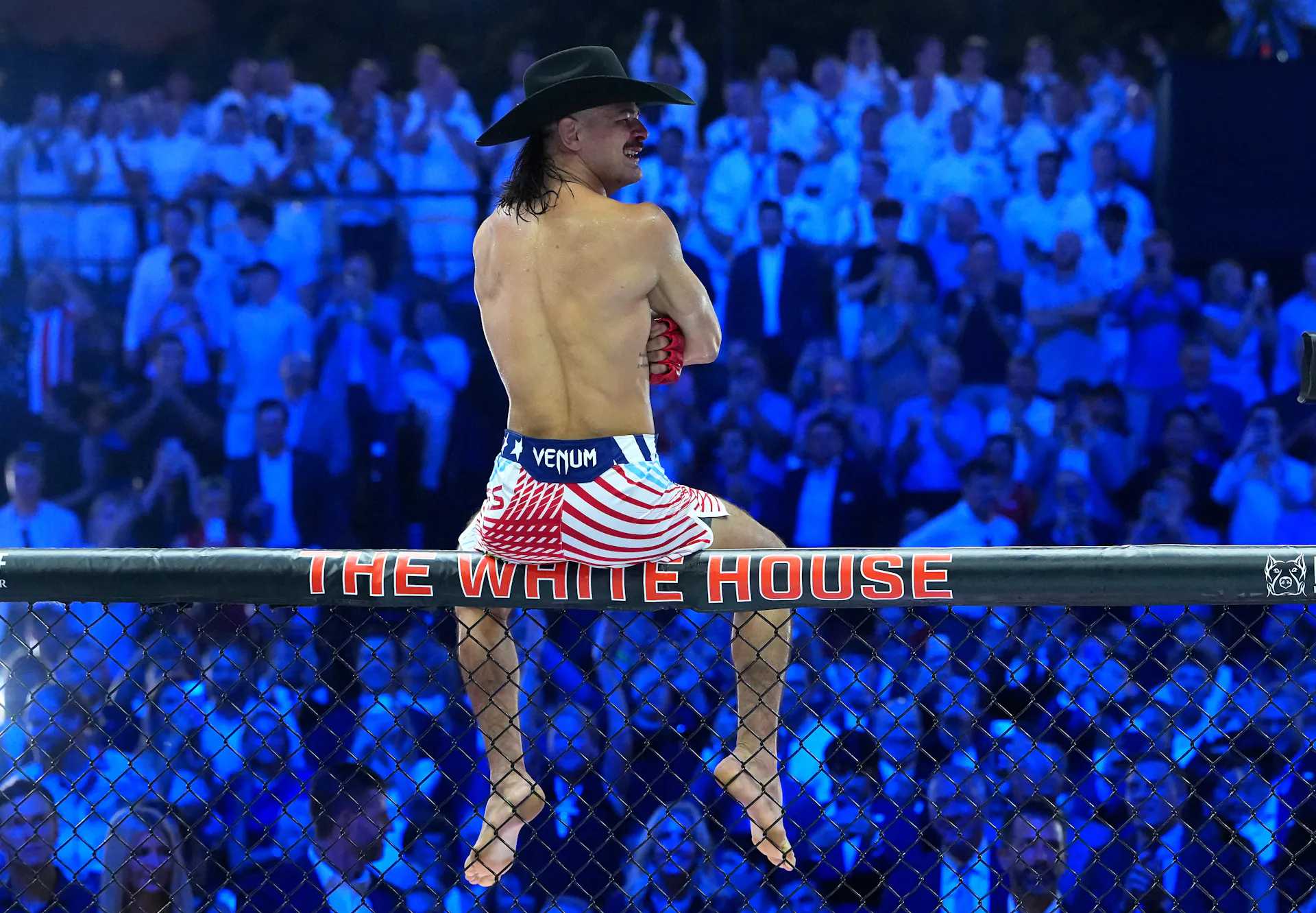 A man in U.S.-flag themed shorts and no shirt sits atop a fence that says 'The White House' before a large crowd in the dark.