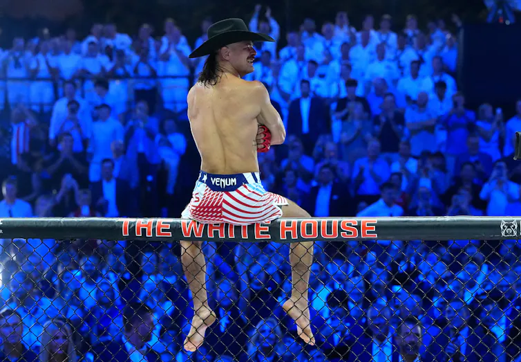 A man in U.S.-flag themed shorts and no shirt sits atop a fence that says 'The White House' before a large crowd in the dark.