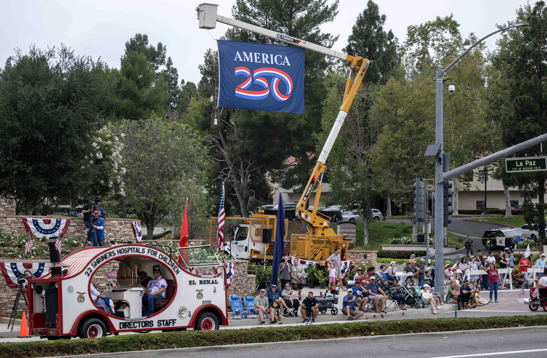 An America250 banner flies overhead at a parade with US flag themed motifs.