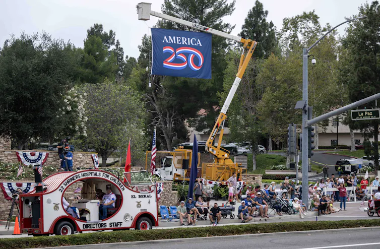 An America250 banner flies overhead at a parade with US flag themed motifs.