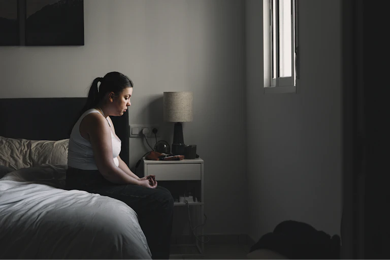 A young woman sits on her bed looking dejected