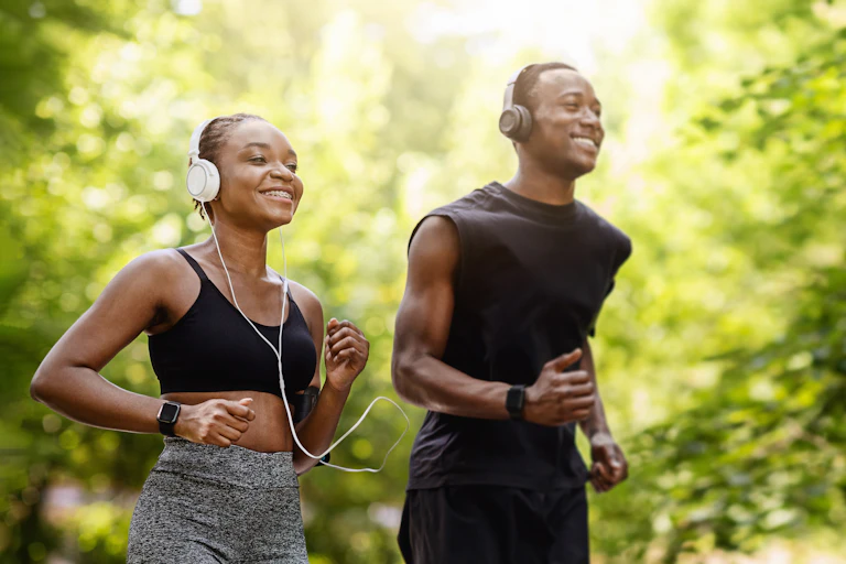 A couple goes for a run outdoors in the woods. They're both smiling.
