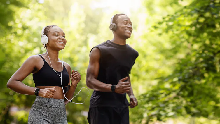 A couple goes for a run outdoors in the woods. They're both smiling.