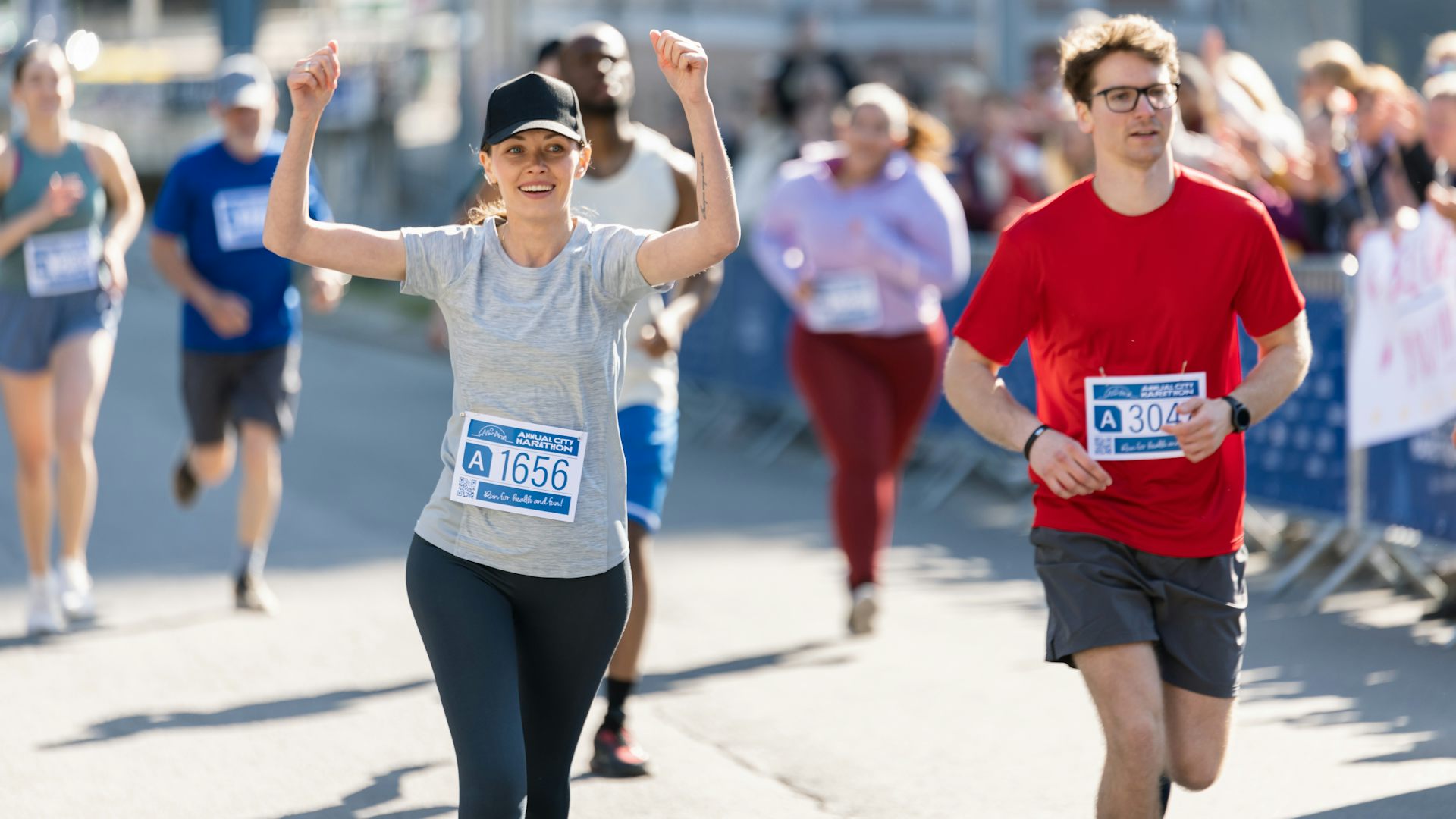 A group of marathon runners wearing race bibs attached to their shirts. A women, who is in the foreground, raises her hands in triumph while smiling.