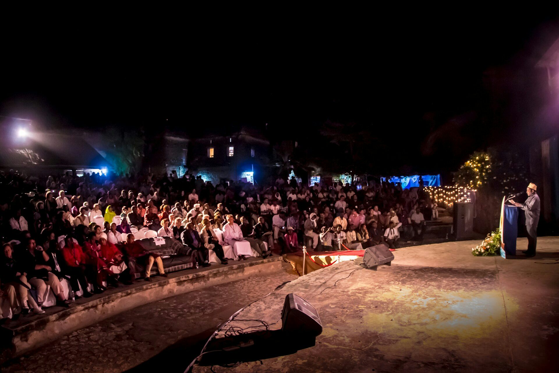 A wide shot of an open air arena. A man stands on a round stahe at a lectern. speaking to a full audience, fairy lights and blue lights behind them.