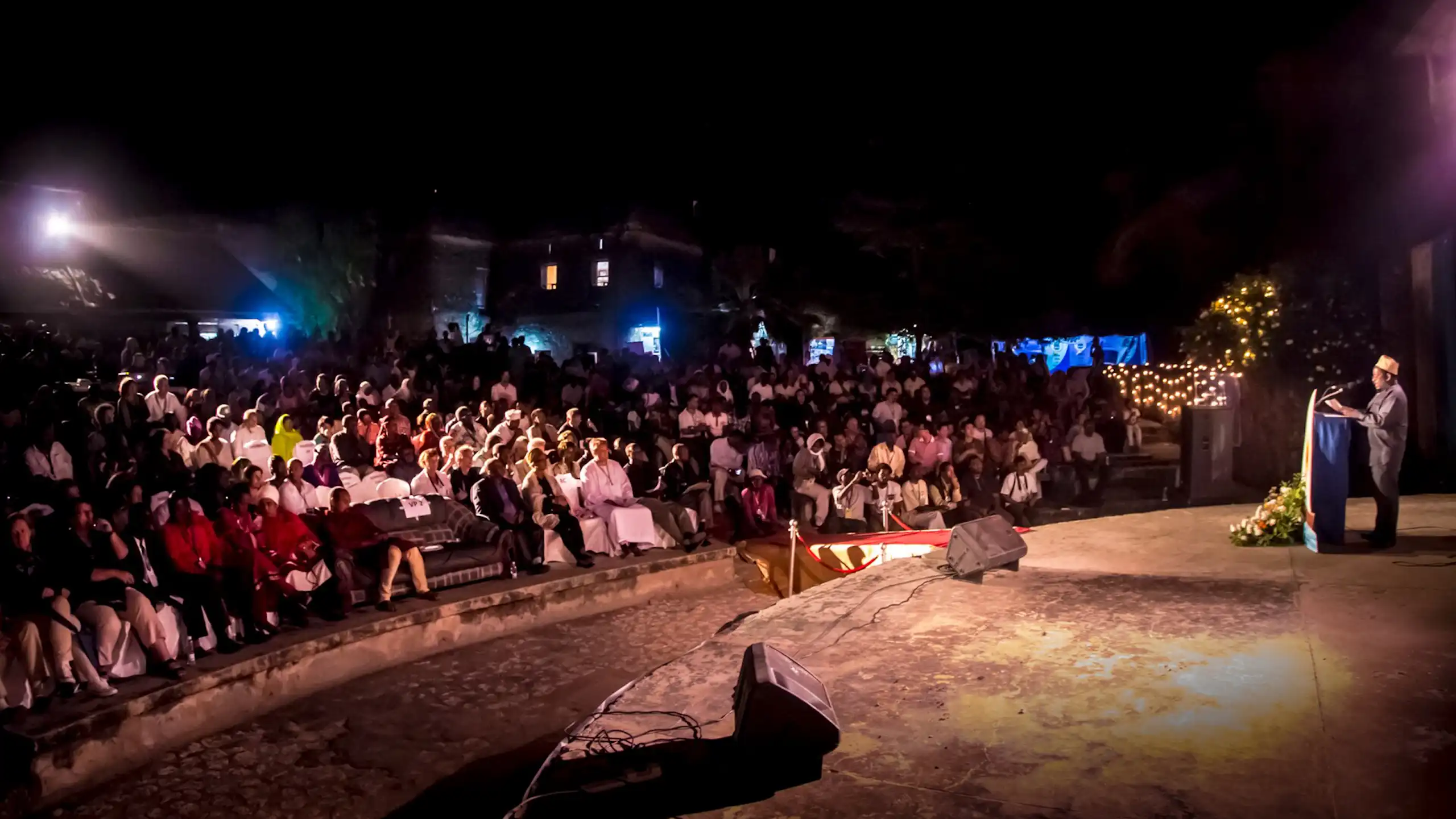 A wide shot of an open air arena. A man stands on a round stahe at a lectern. speaking to a full audience, fairy lights and blue lights behind them.