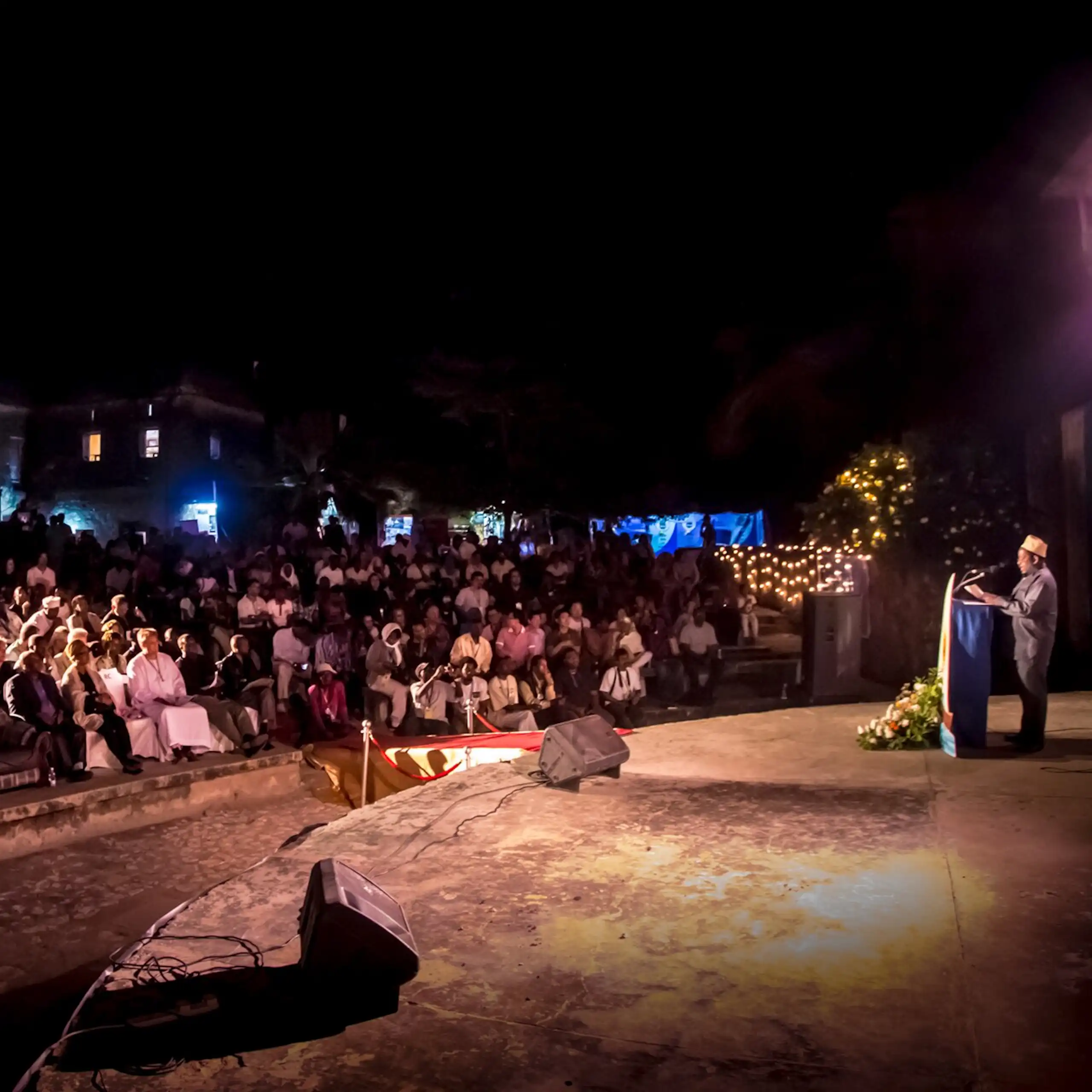 A wide shot of an open air arena. A man stands on a round stahe at a lectern. speaking to a full audience, fairy lights and blue lights behind them.