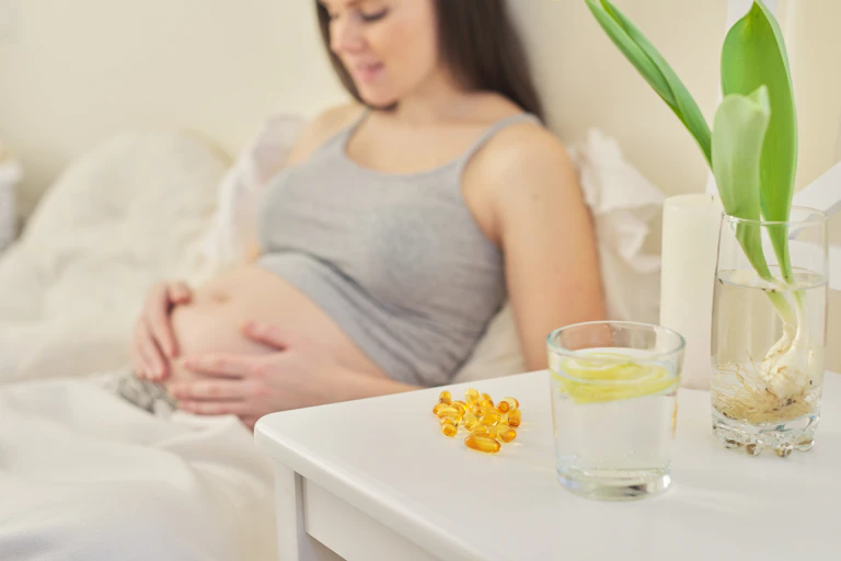 Pregnant woman in bed in background. Vitamin D capsules next to glass of water on bedside table in foreground.