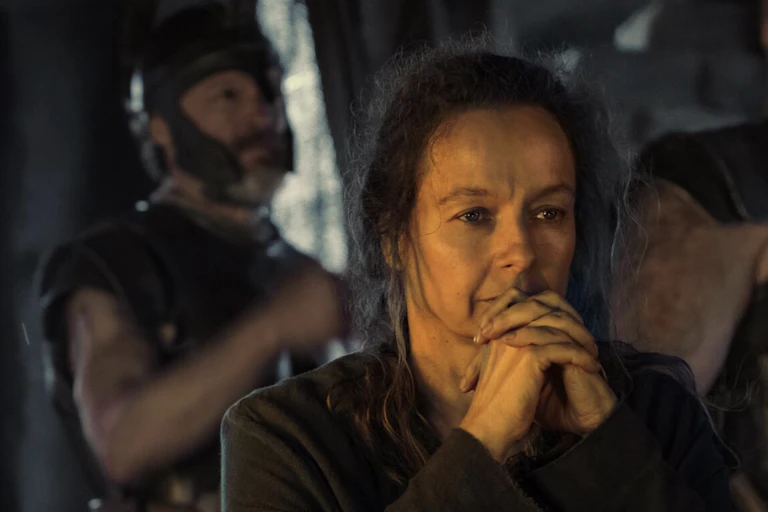A middle-aged woman with messy hair clasps her hands and rests her chin pensively on them.