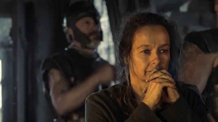 A middle-aged woman with messy hair clasps her hands and rests her chin pensively on them.