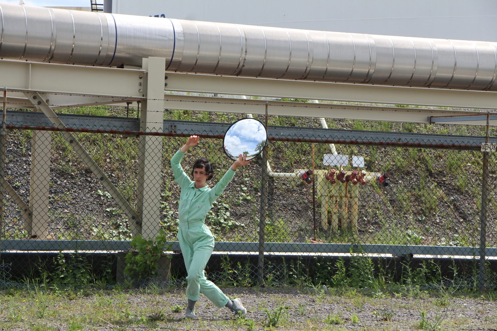 A person in green coveralls lunging to the side holding up a round mirror.