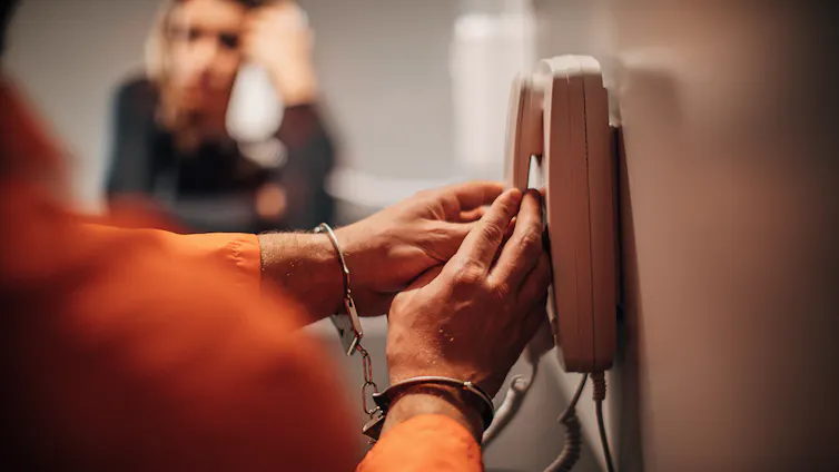 A man with orange sleeves and handcuffs grabs a phone mounted on the wall. A person on the other side of a glass barrier holds a phone to their ear.