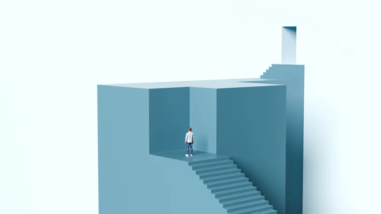 Man standing at the top of a staircase blocked by a solid wall looking toward a door located above him.