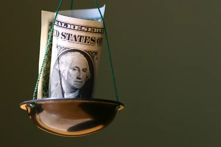 A folded U.S. dollar bill sits in one side of a balance scale.