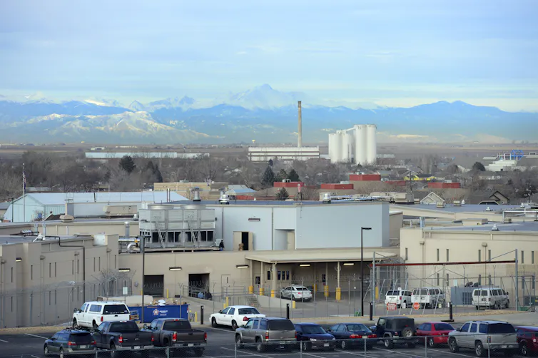 A tan building is fenced in by a wire fence. Mountains are in the background.