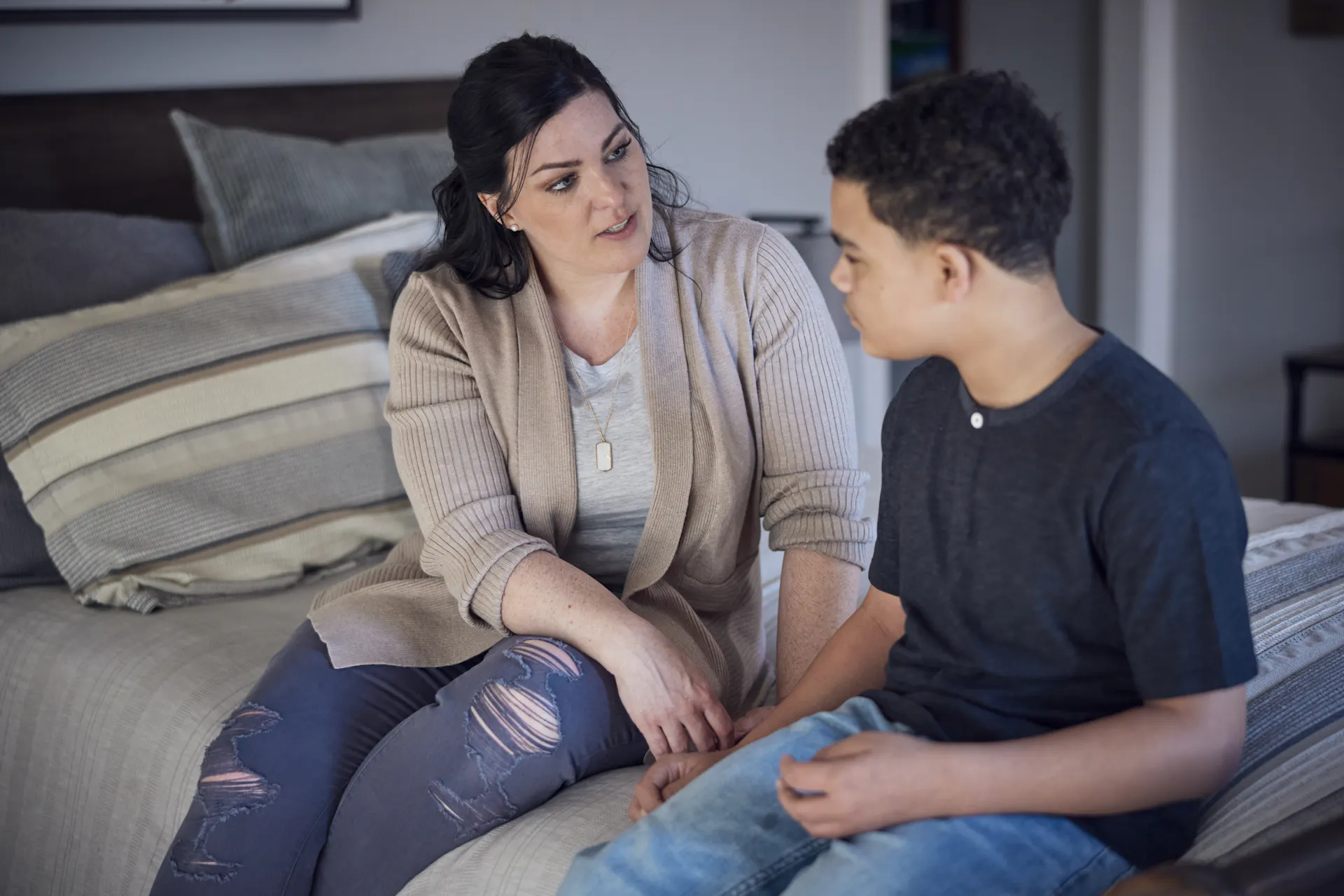 A middle-aged woman with a serious expression sits on a bed talking with a young boy.