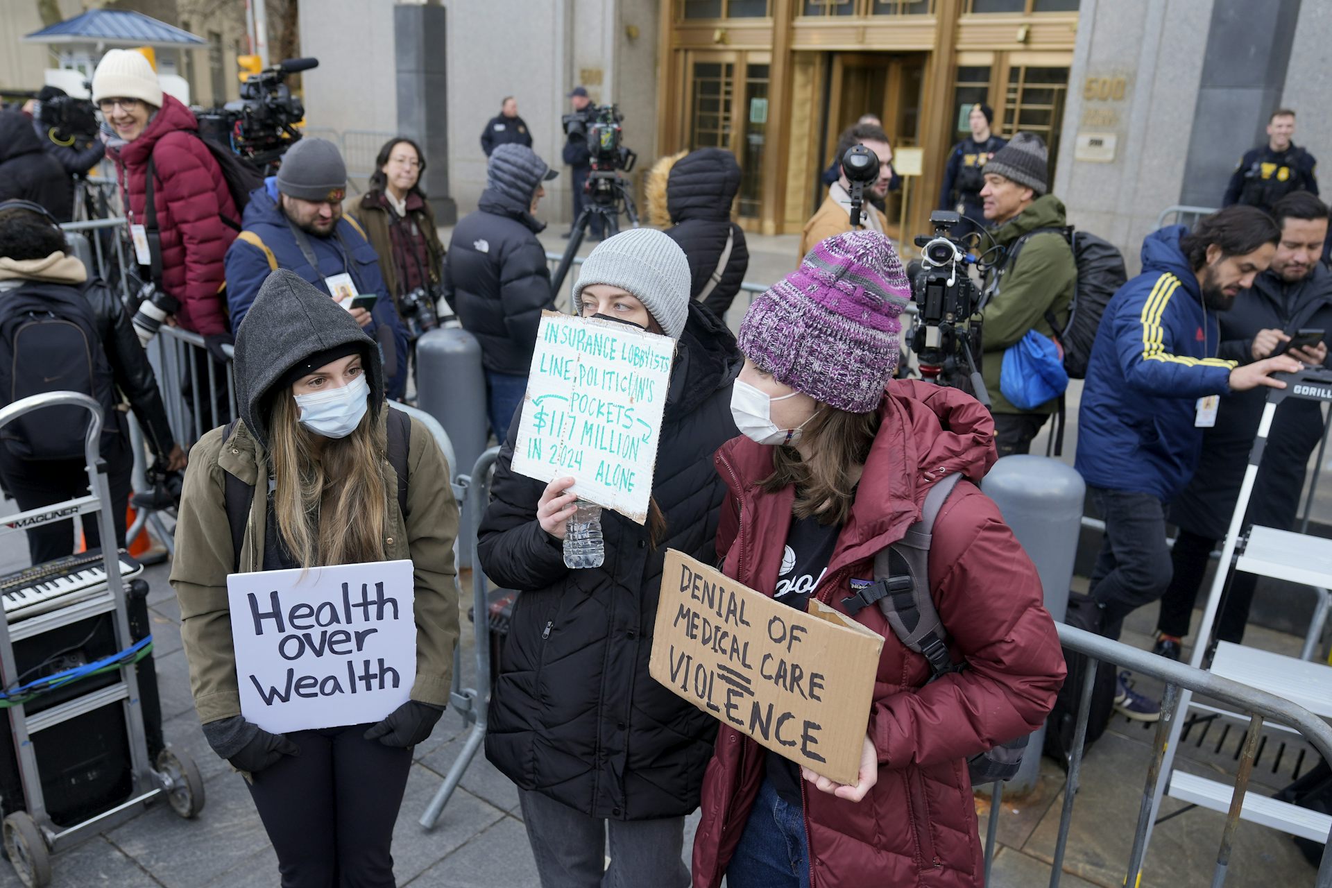 Demonstrators wearing face masks stand outside court holding signs that read 'health over wealth' and 'denial of medical care = violence'
