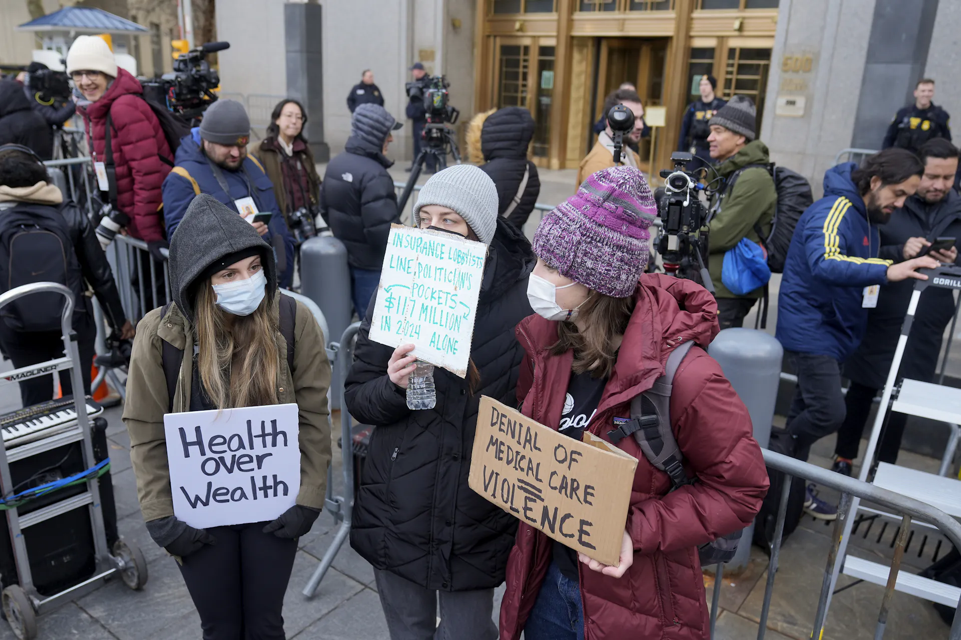Demonstrators wearing face masks stand outside court holding signs that read 'health over wealth' and 'denial of medical care = violence'