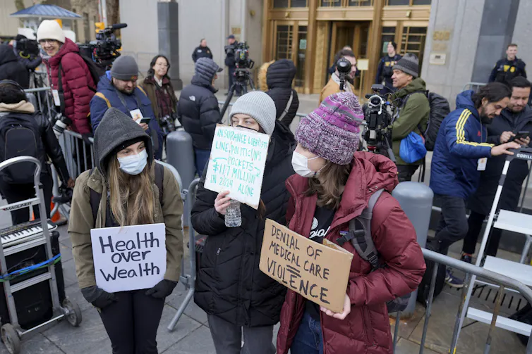 Demonstrators wearing face masks stand outside court holding signs that read 'health over wealth' and 'denial of medical care = violence'