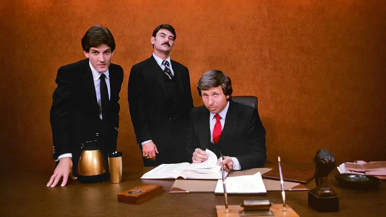 Three businessmen in suits stare intently into the camera from behind a large desk, with coffee, documents and pens