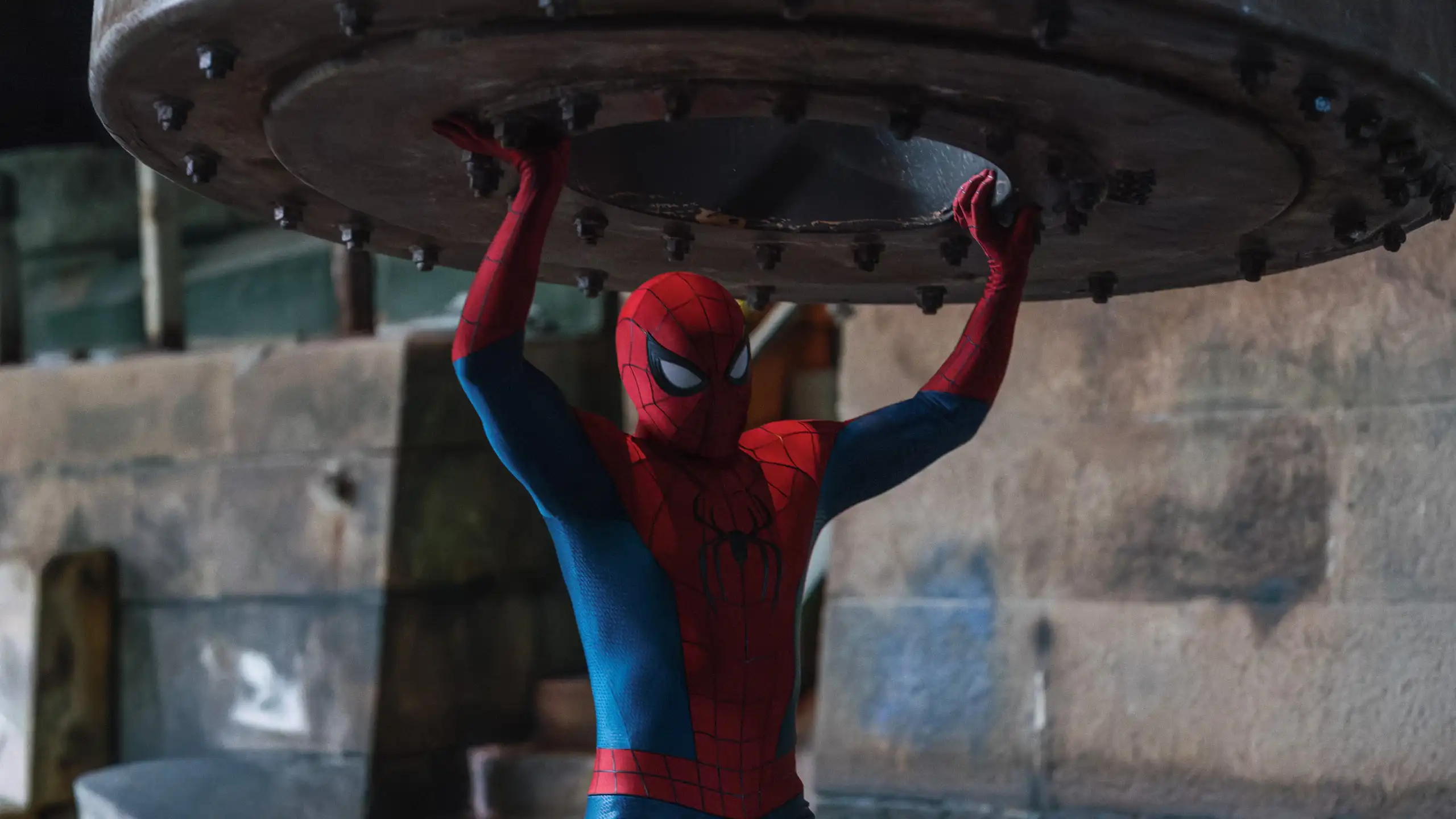 Spider-Man holds a large metal object overhead.