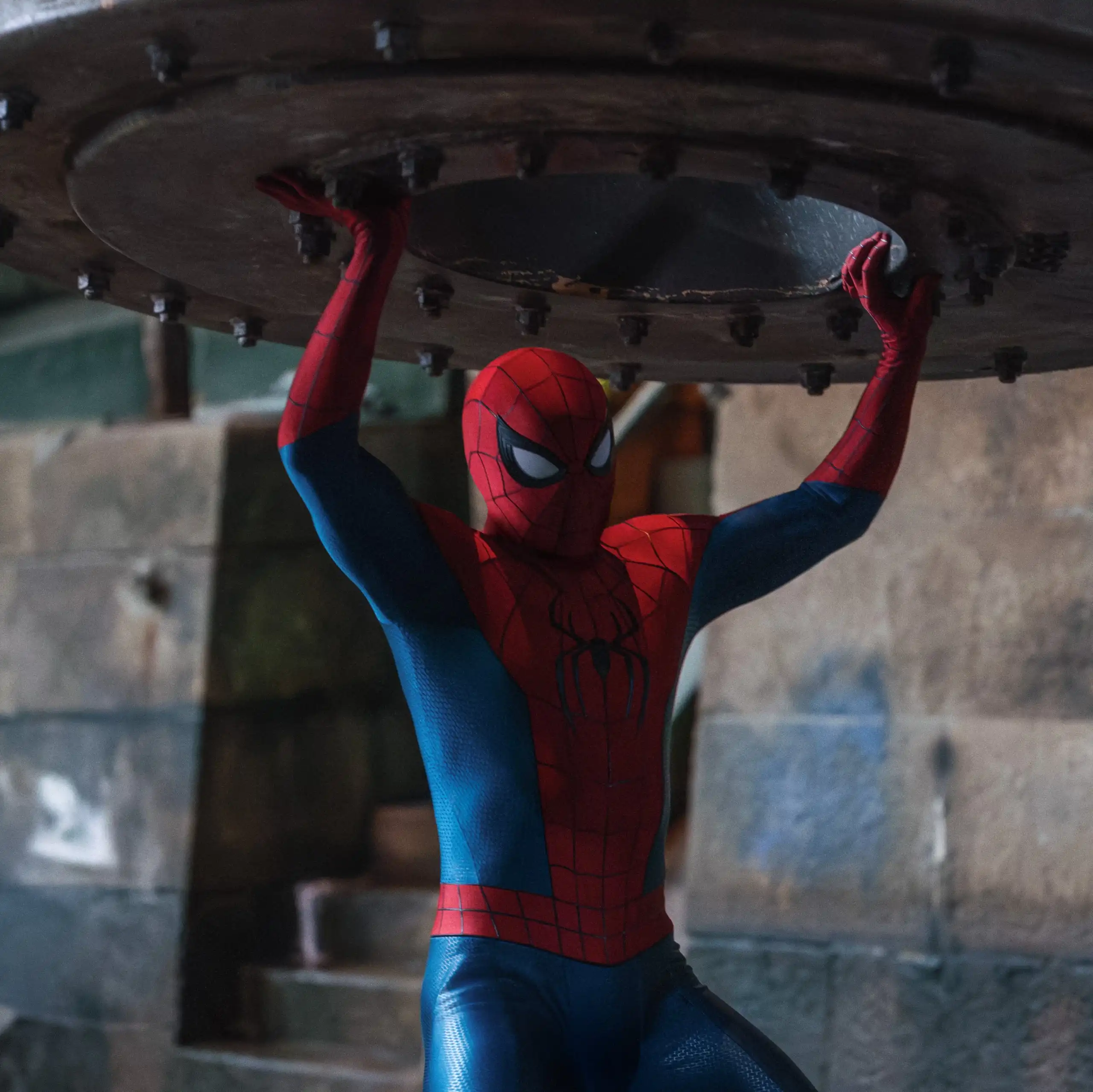Spider-Man holds a large metal object overhead.