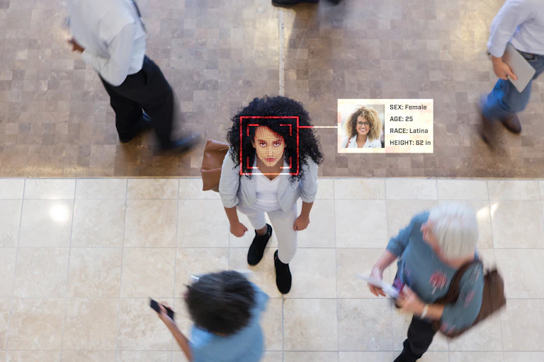 A young businesswoman looks up while in an office lobby with business people all around her. A facial recognition scan reveal her personal data.