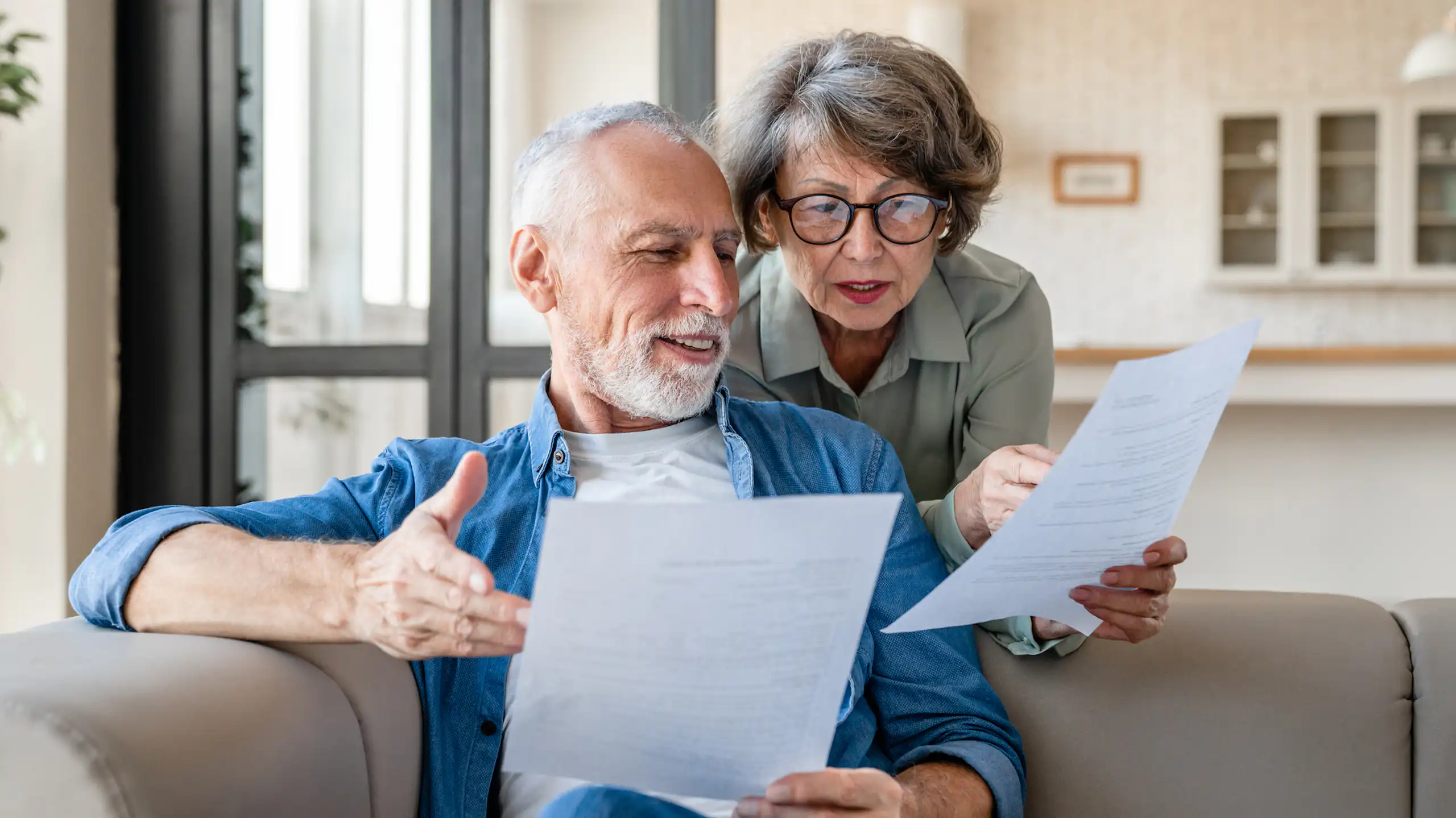 Older man sitting on sofa with paperwork, older woman standing behind him, holding paperwork