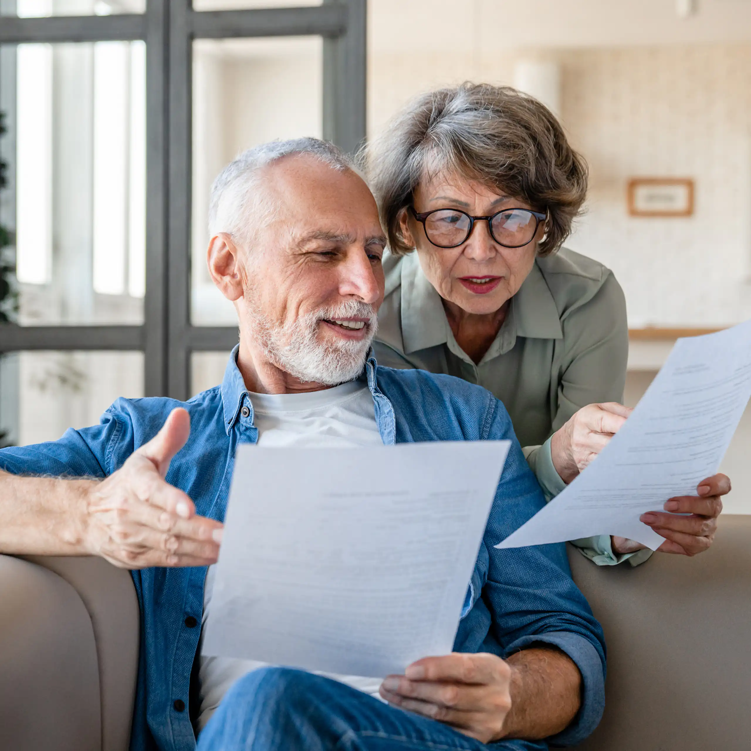Older man sitting on sofa with paperwork, older woman standing behind him, holding paperwork
