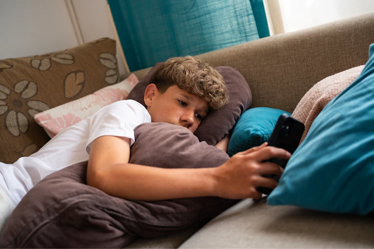 A teenage boy is relaxed on a couch, surrounded by soft cushions, focused on his smartphone.