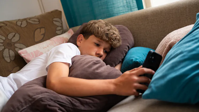 A teenage boy is relaxed on a couch, surrounded by soft cushions, focused on his smartphone.