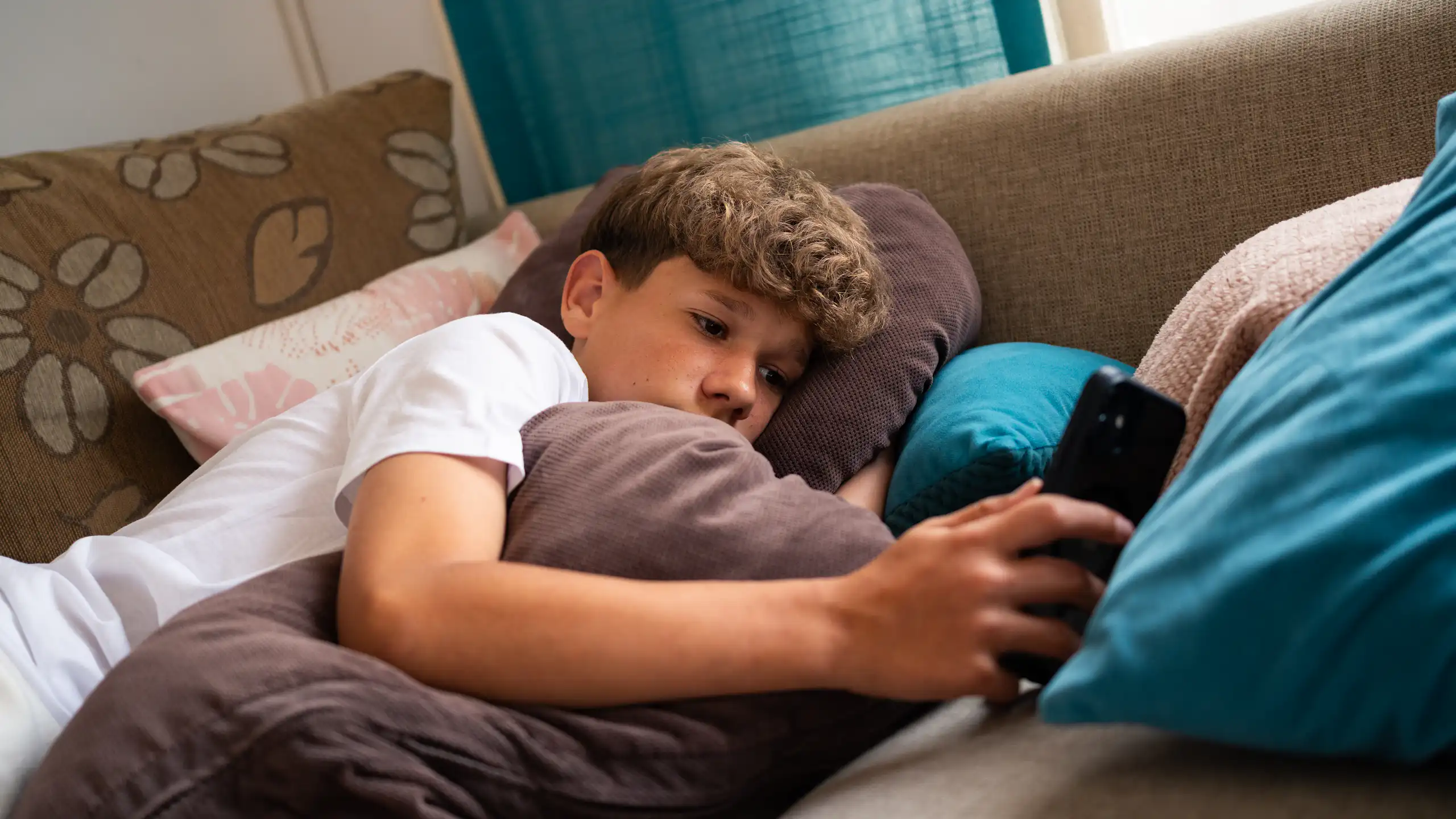 A teenage boy is relaxed on a couch, surrounded by soft cushions, focused on his smartphone.